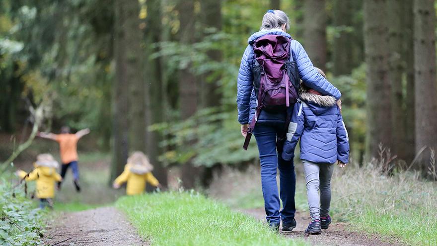 Mom hiking in forest with her arm on young girls shoulders with rest of family hiking ahead.