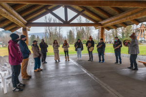 a group people standing under a log roof singing and drumming